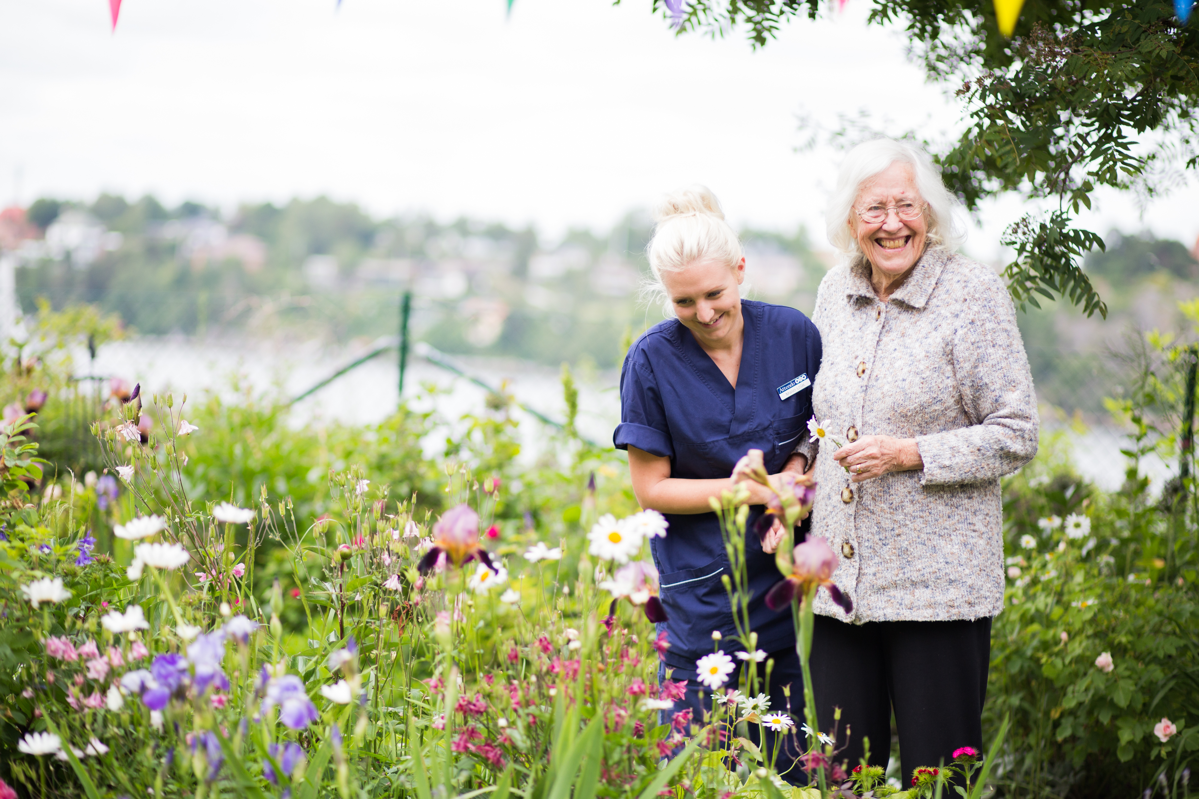 Sjuksköterska och äldre kvinna njuter av trädgårdsarbete bland blommor, med en sjö i bakgrunden