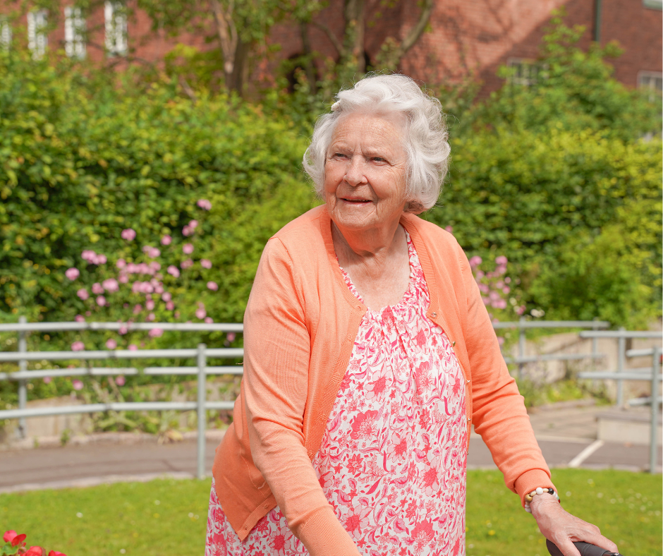 Äldre dam i färgglada kläder står med sin rullator och blickar ut över en grönskande trädgård