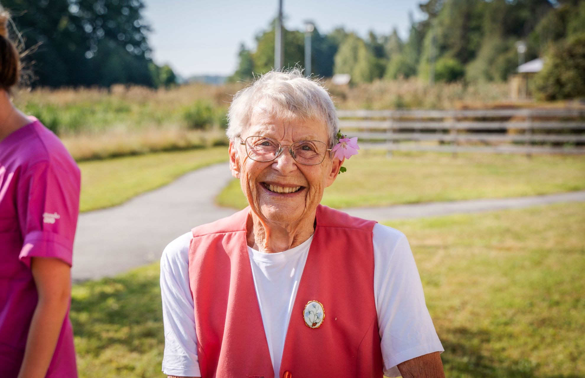 Äldre kvinna med blomma bakom örat ler stort, bredvid personal från Attendo i grön omgivning
