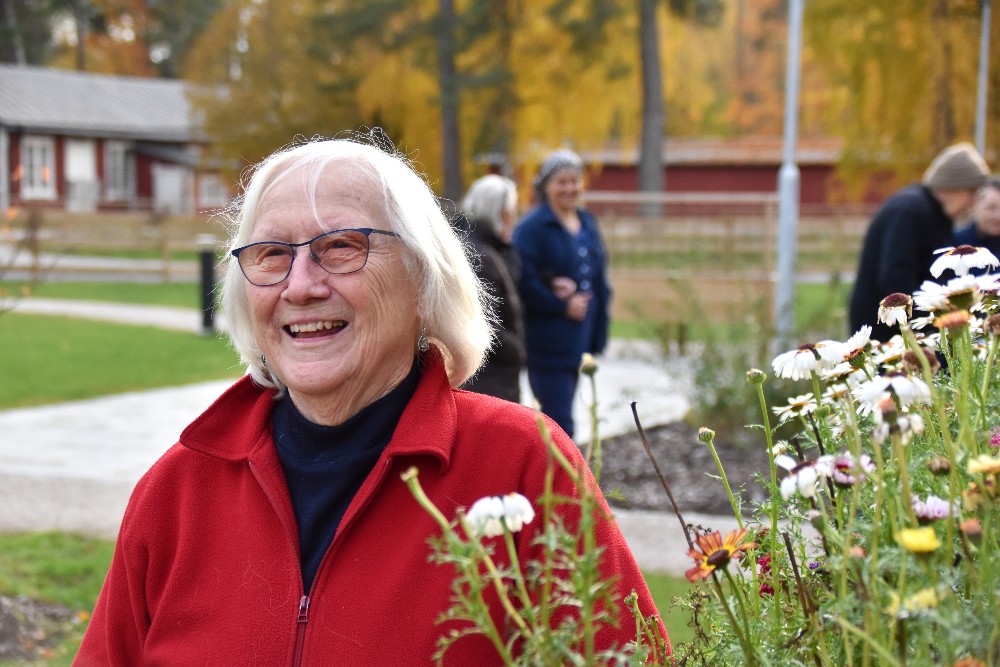Leende äldre kvinna med glasögon och röd jacka vid blommor i en park, med personer och höstträd i bakgrunden