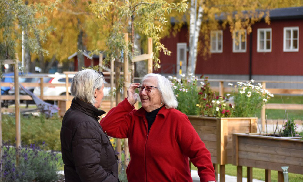 Två äldre personer samtalar utomhus vid en trädgård med höstliga färger och blommor