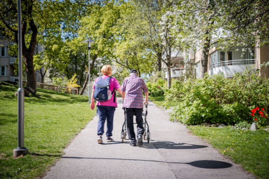 Hemtjänstkund med rollator och personal promenerar tillsammans i grönska