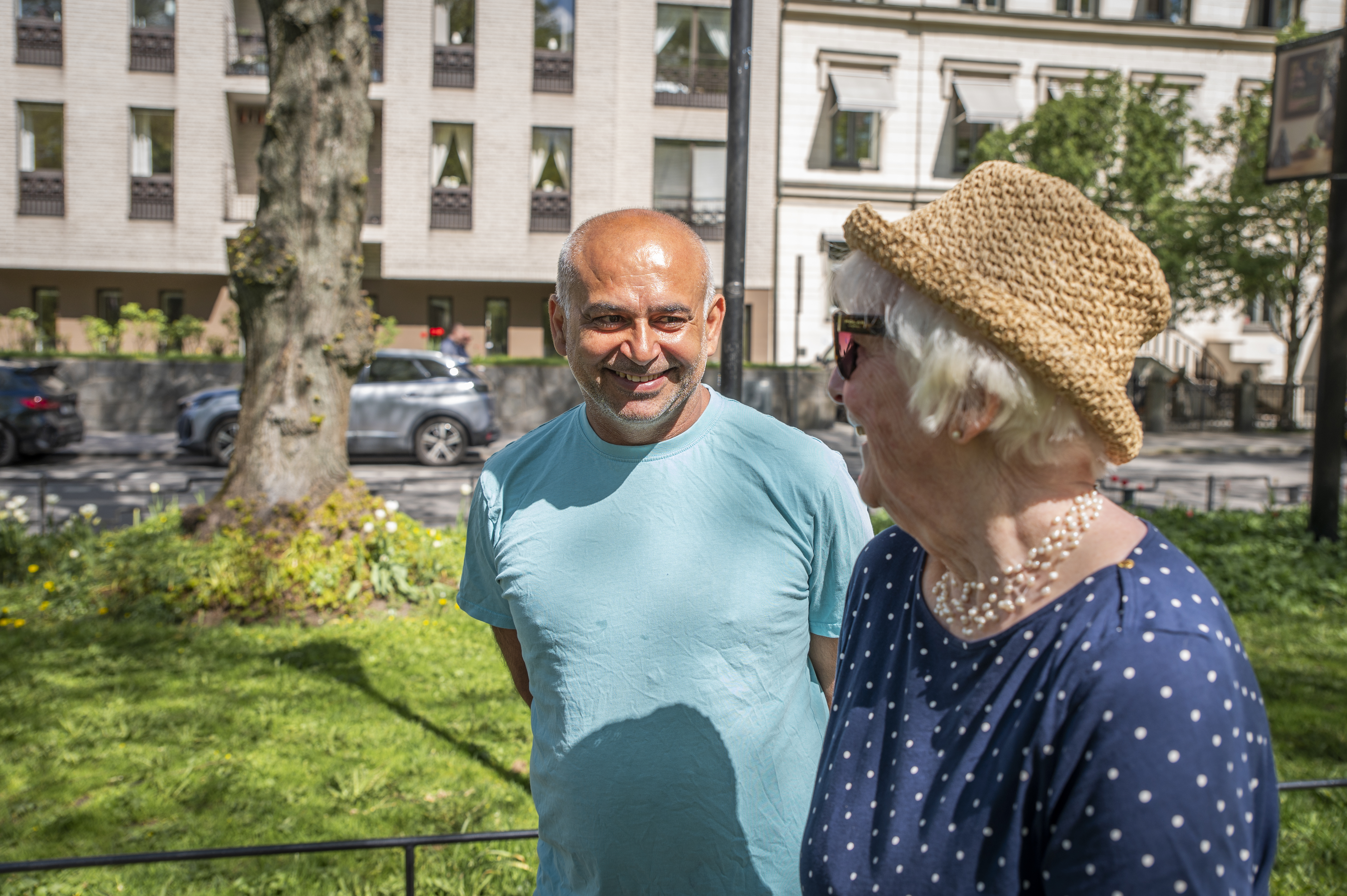 Två personer står och pratar i en grön parkmiljö med träd, gräsmatta och hus i bakgrunden. Utebild i solsken